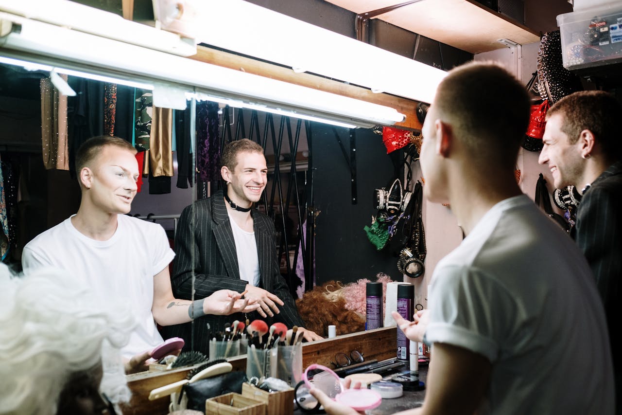 Two men in a dressing room preparing makeup, reflected in a mirror, showcasing transformation and identity.