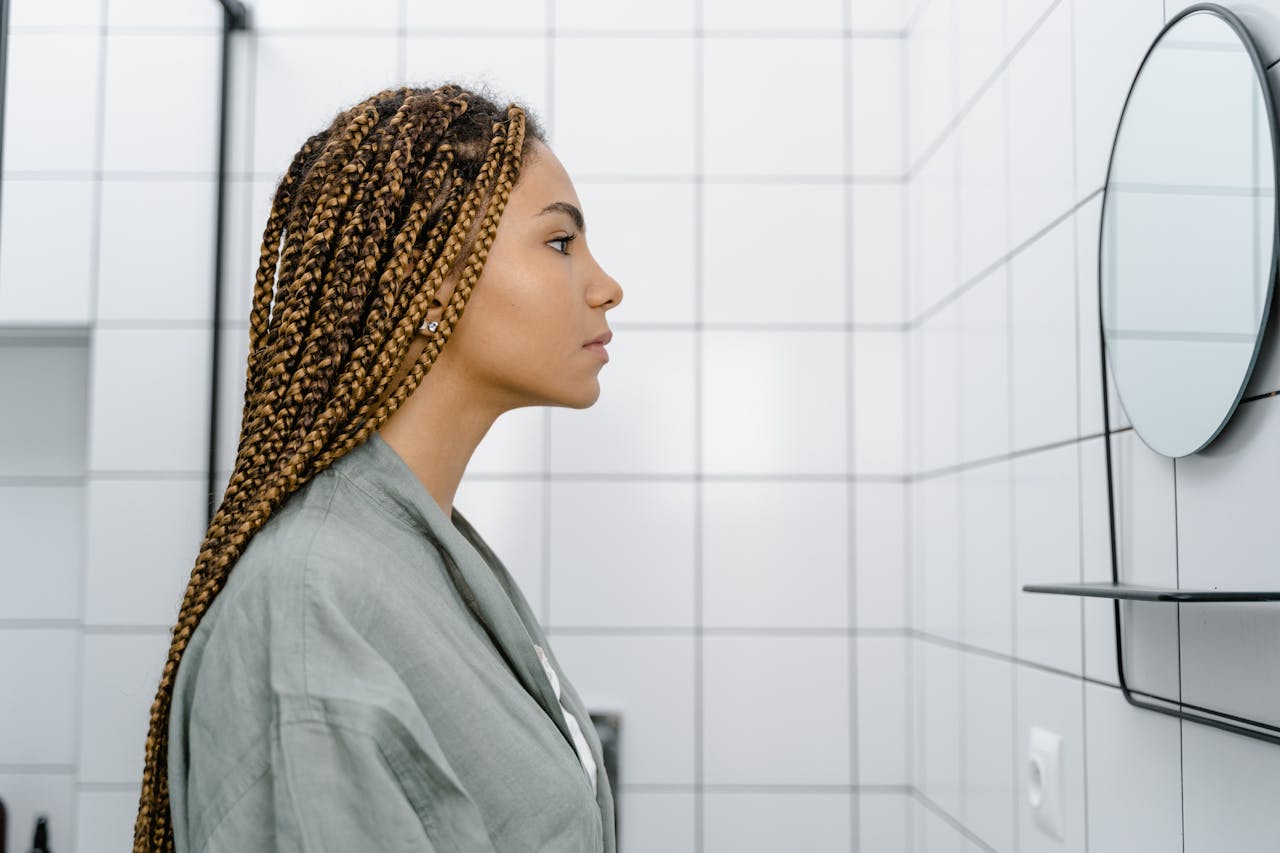Profile view of a woman with braided hair looking at her reflection in a modern tiled bathroom.